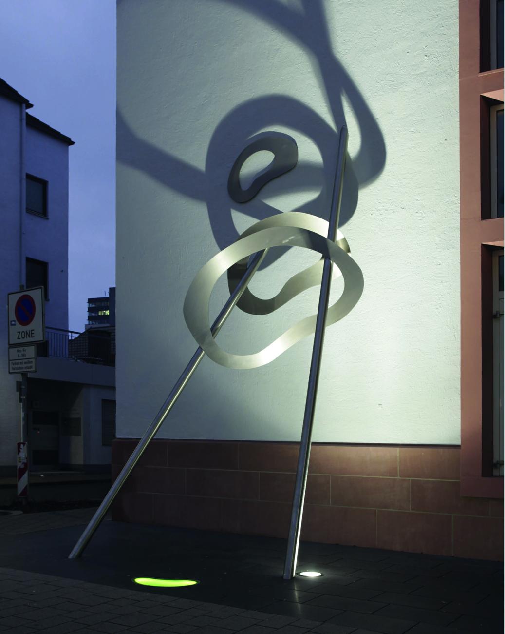 Am Abend lässt der Strahler die Formen als große Schatten über die weiße Fassade wandern und verwandelt den Vorplatz.
